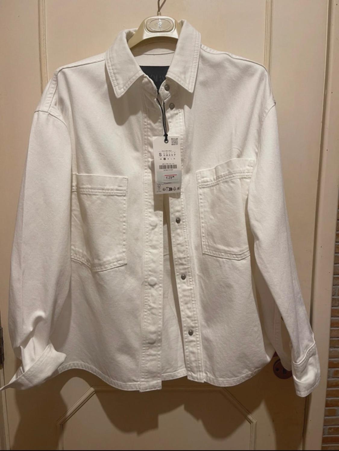 White long-sleeved shirt with front pockets, hanging on a hanger. Visible label with size and price details.