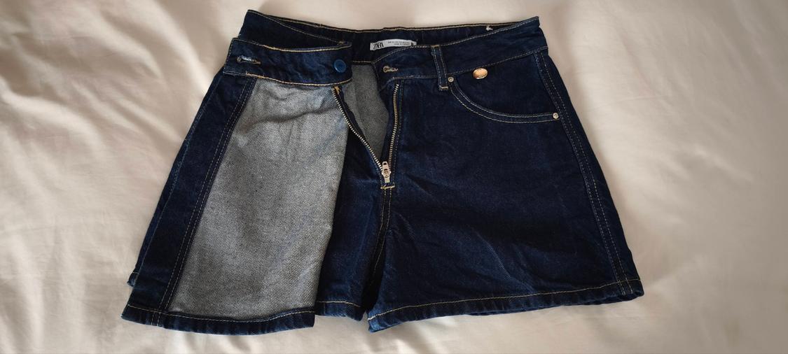 Dark blue denim shorts with a grey inner panel, front zip, and a gold metallic button on a white background.