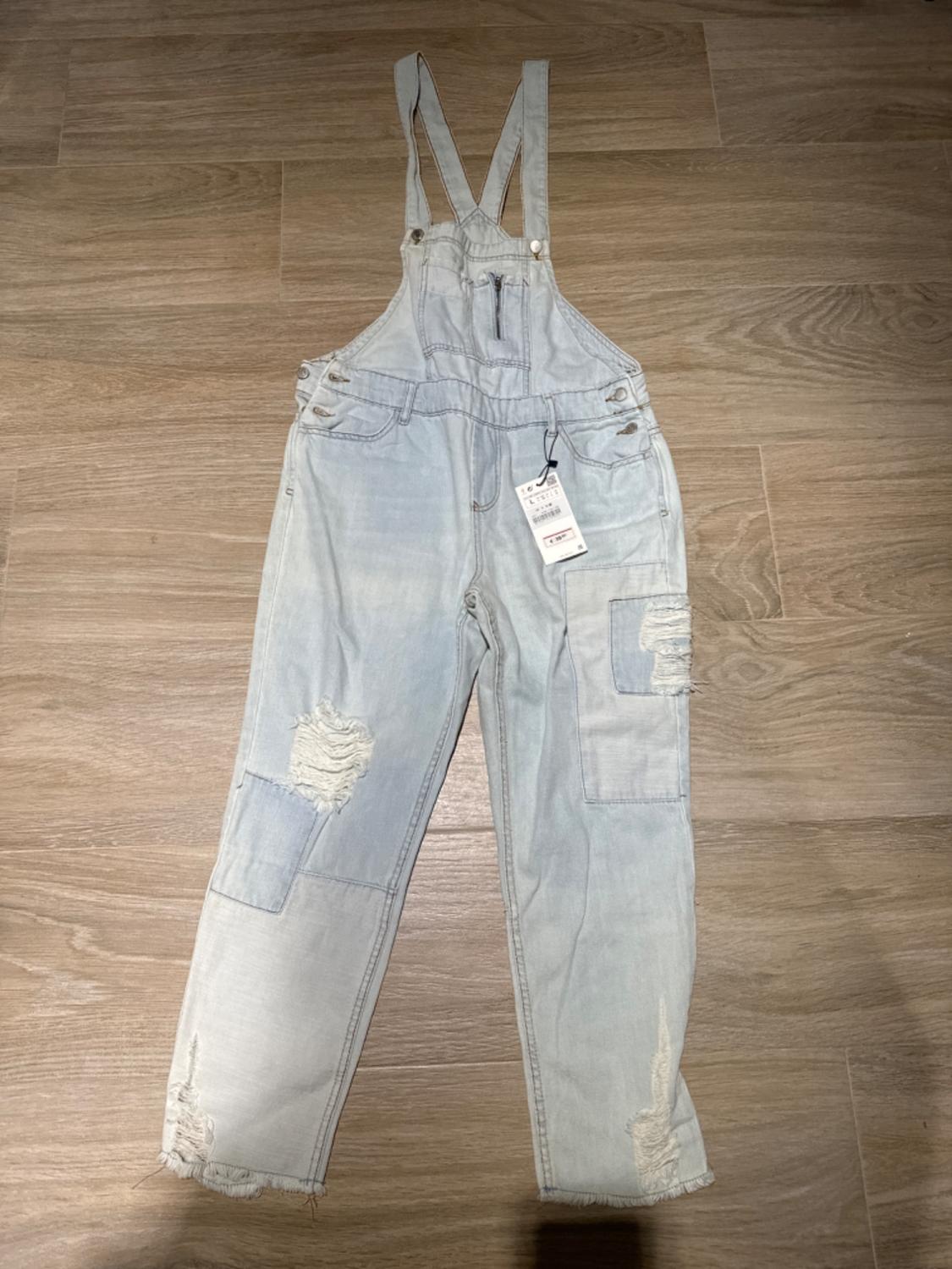 Front view of light blue denim overalls with straps, pockets and distressed details.