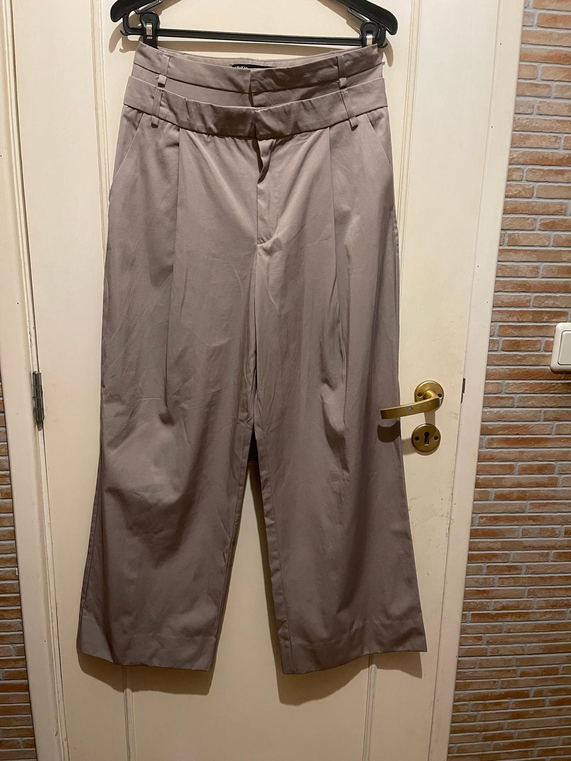Wide-leg beige trousers hanging on a black hanger over a white door, with a brick wall background.