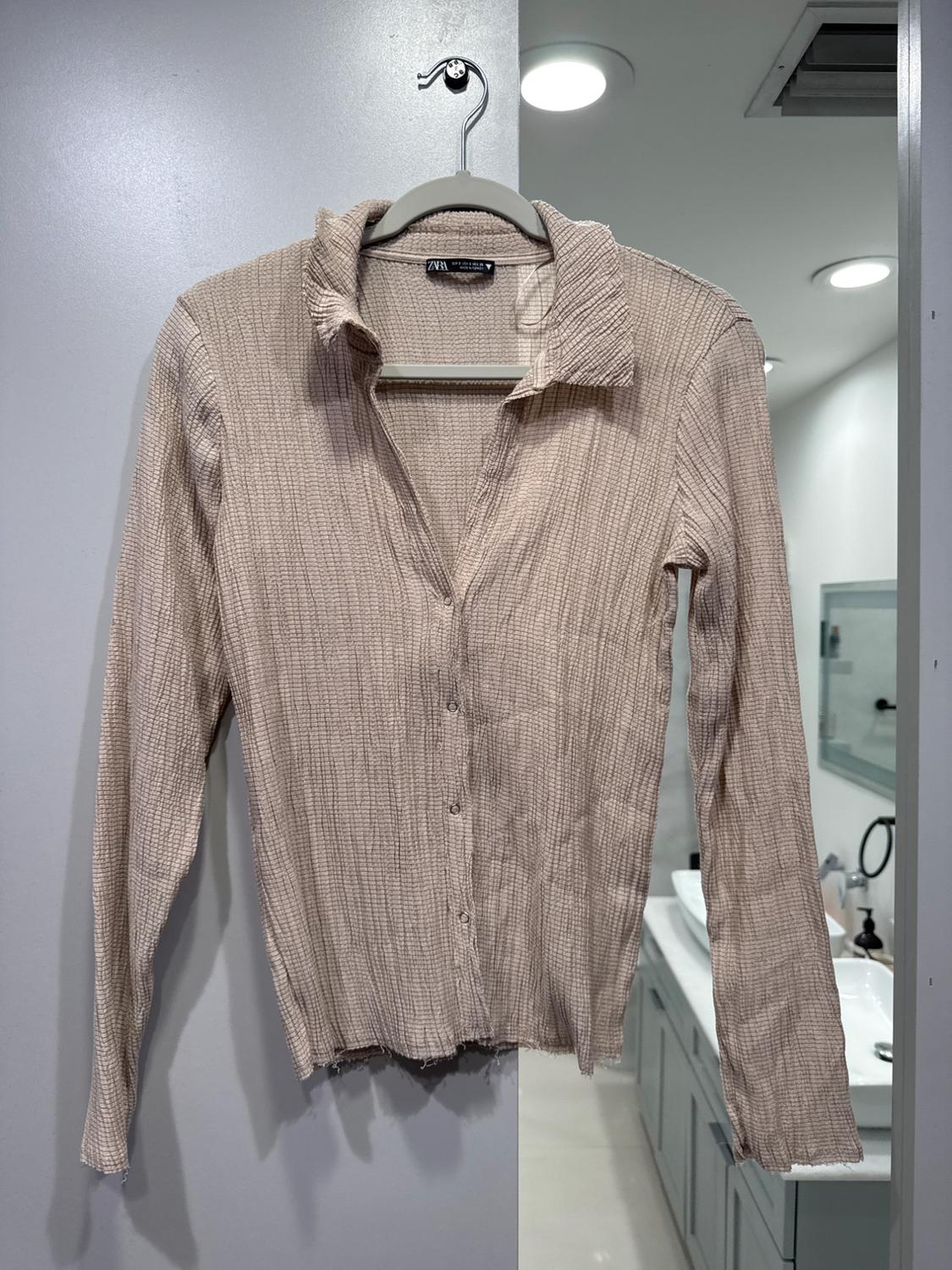 Beige long-sleeleeved shirt with wrinkled texture, hanging on a hanger in a modern bathroom with ceiling lights.