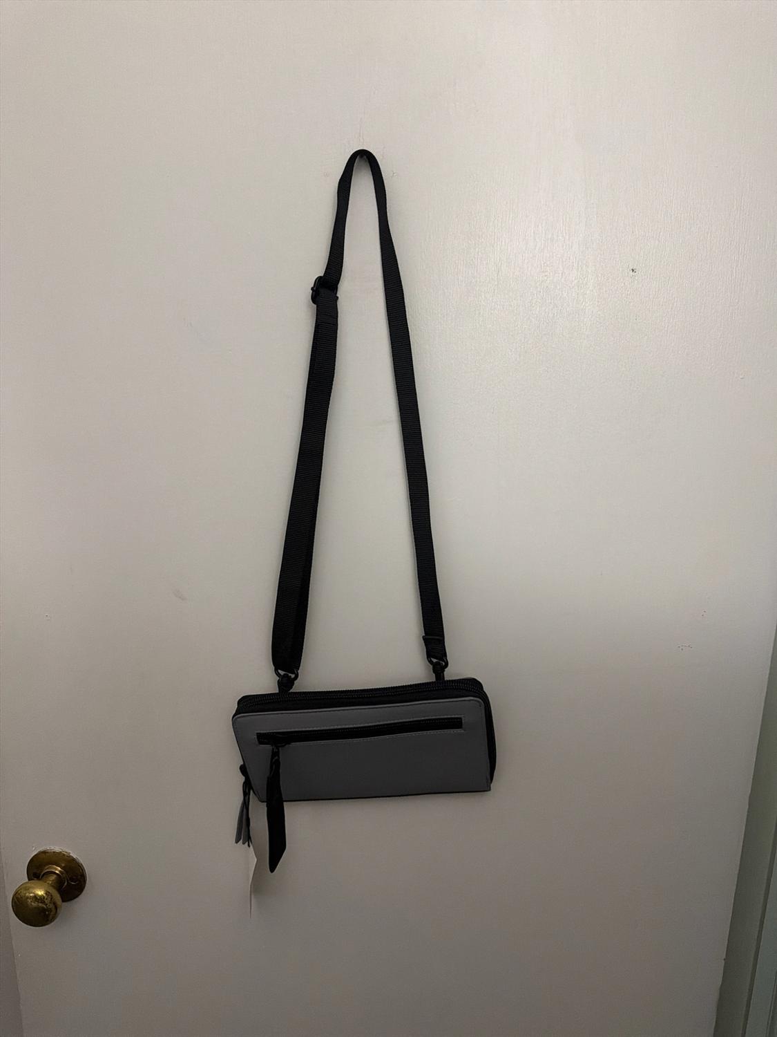 Gray rectangular crossbody bag with a black strap hanging on a white door with a golden knob.