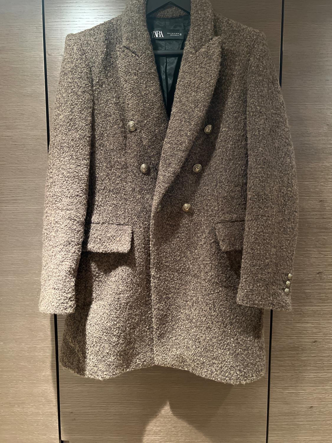 Textured brown coat with gold buttons, hanging on a black hanger against a light wooden background. Label visible inside.