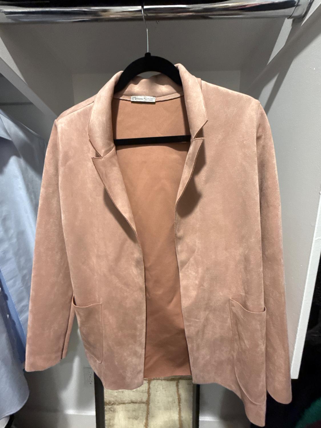 Powder pink blazer-style jacket with lapels and pockets, hanging on a black hanger inside a closet.