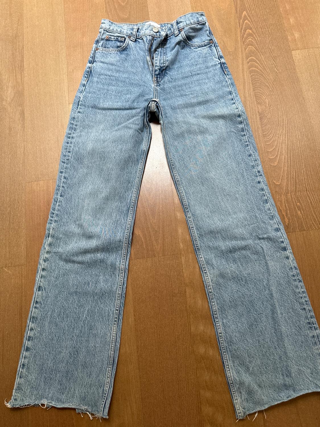 Light blue denim jeans with a straight cut and frayed hem, placed on a brown wooden floor.
