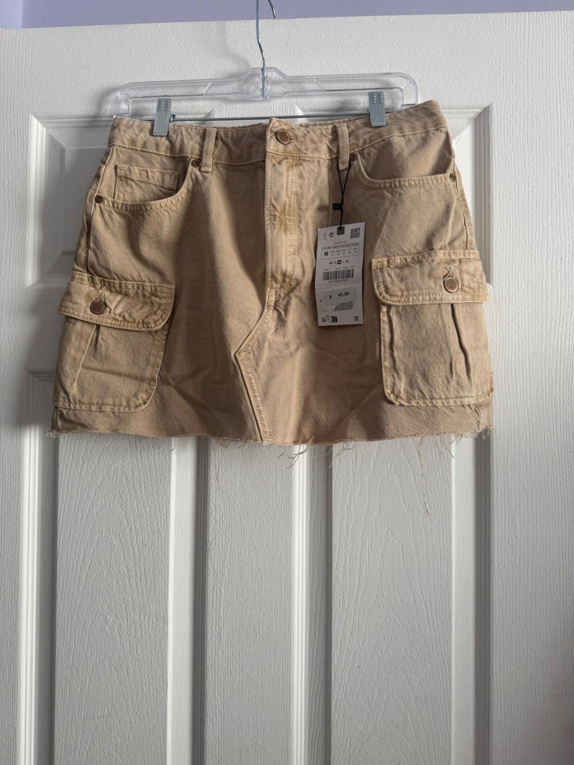 Short beige skirt with large pockets and a swing tag, hanging on a clear hanger over a white door.