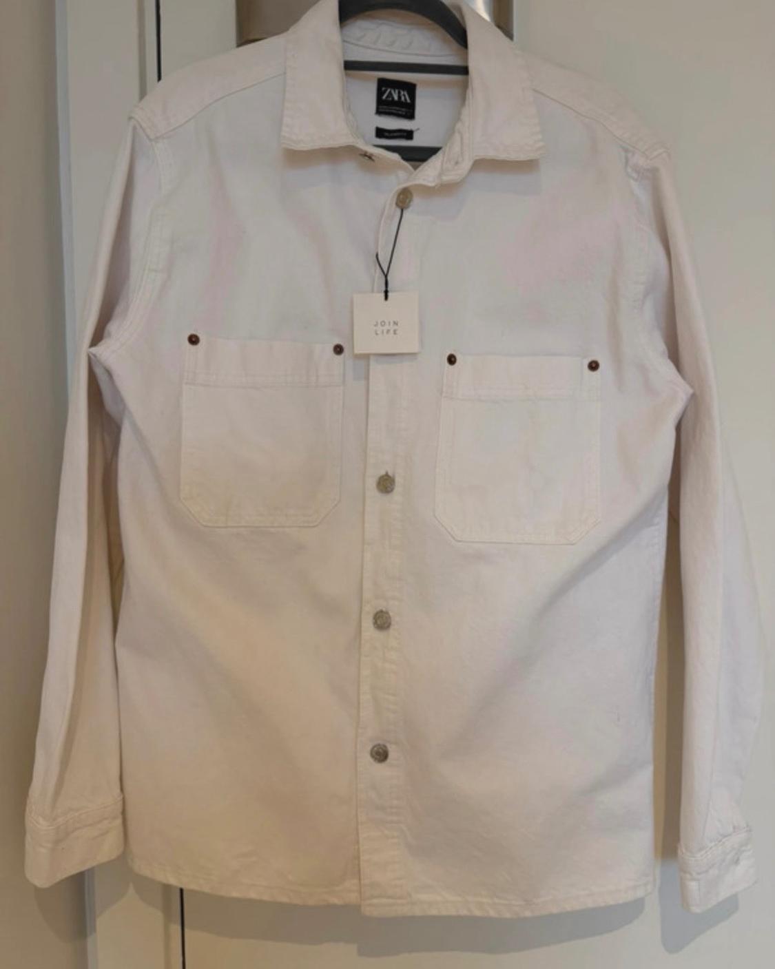 Long-sleeved white button-up shirt hanging on a hanger. Two chest pockets. Labels with "ZARA" and "JOIN LIFE" text.