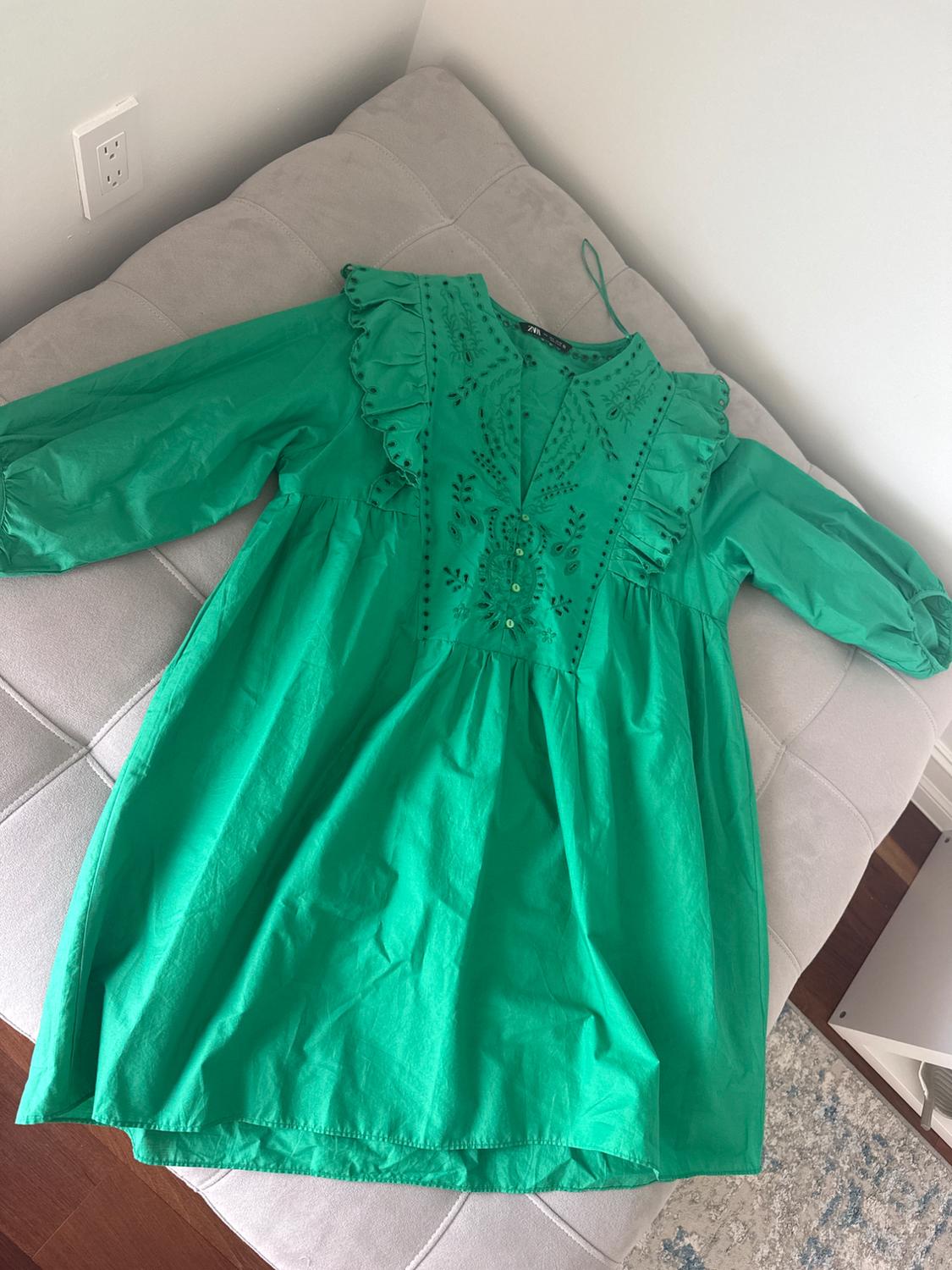 Green dress with ruffles and floral embroidery on the chest, laid out on a light grey sofa.