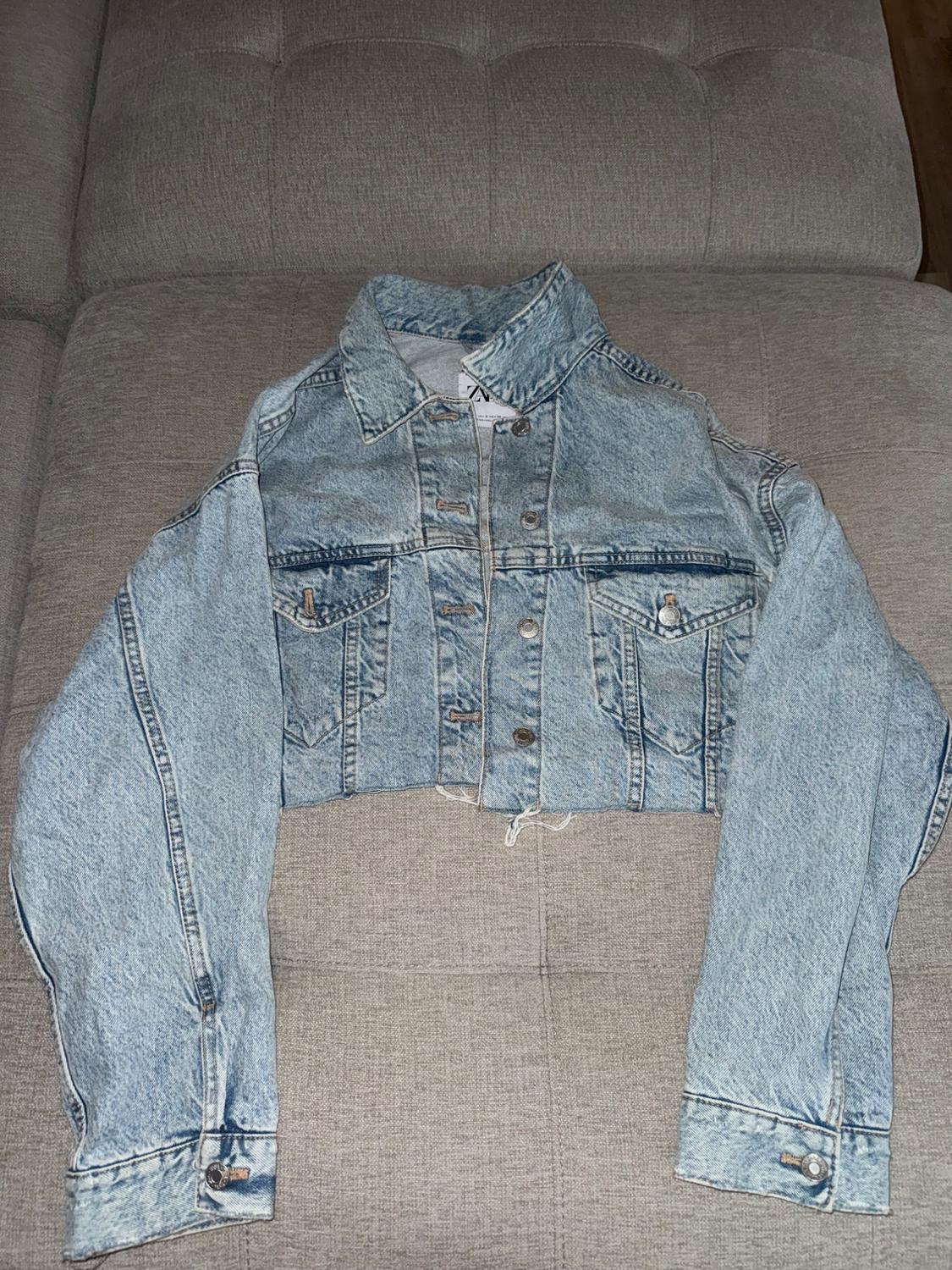 Light blue denim crop jacket with metal buttons and front pockets, placed on a gray sofa.
