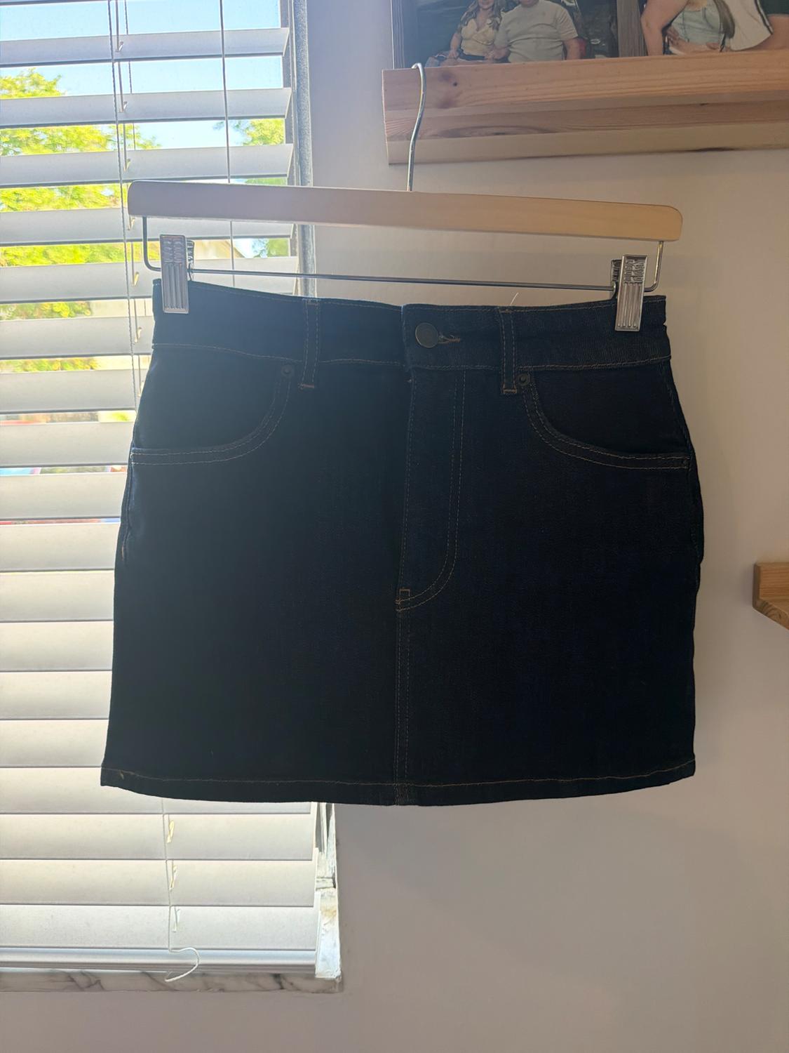 Dark blue denim miniskirt hanging on a wooden hanger in front of a window with white blinds and natural light.