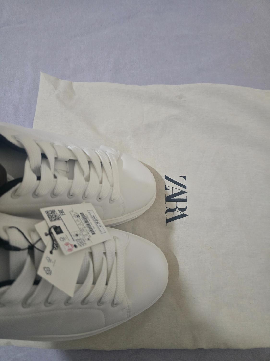 White trainers with laces and tags, resting on a fabric bag bearing the text ZARA. Casual composition.