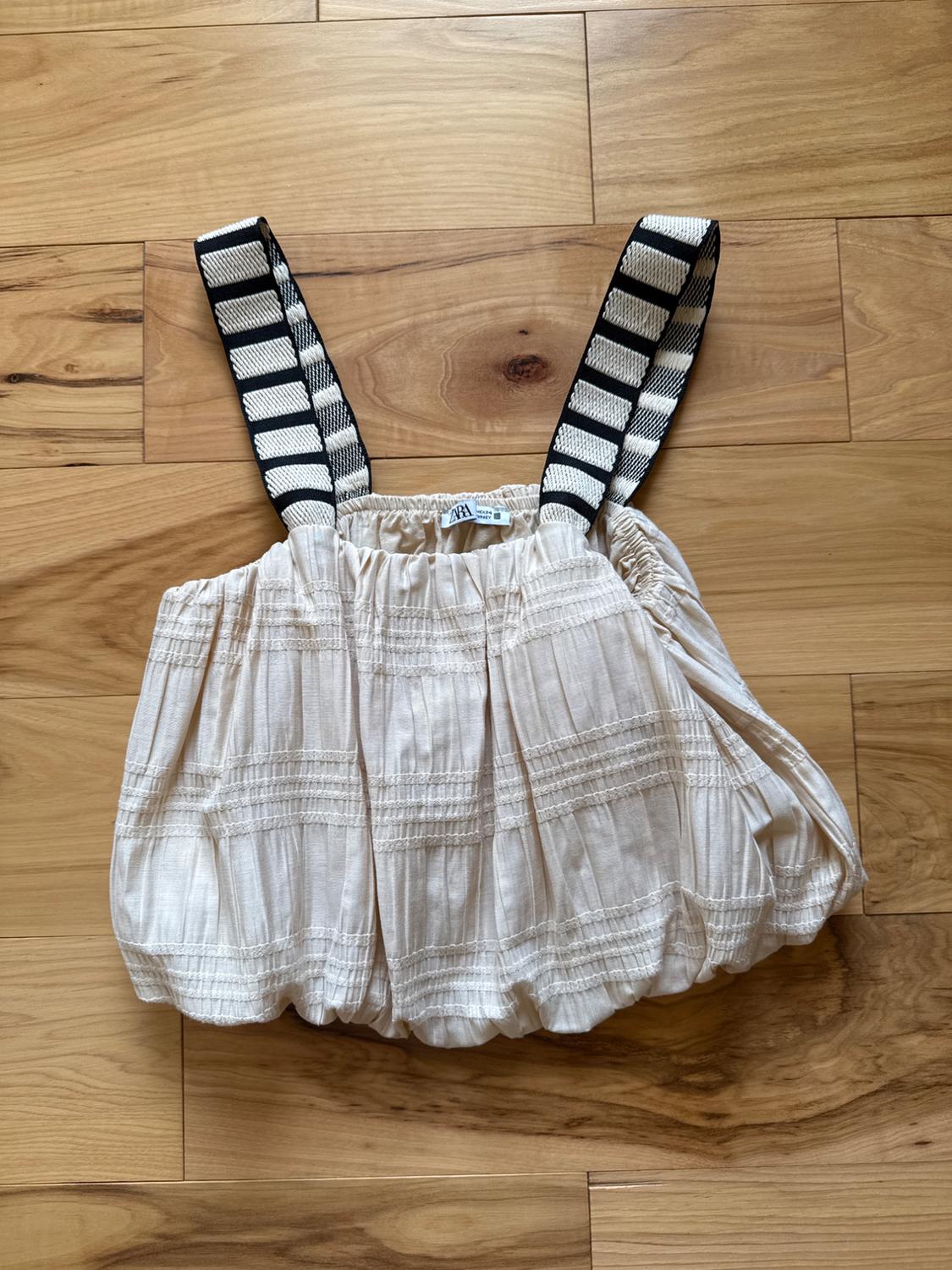 Beige pleated texture top, wide black and white striped straps, on a light wood background. Visible label with "Zara" logo.