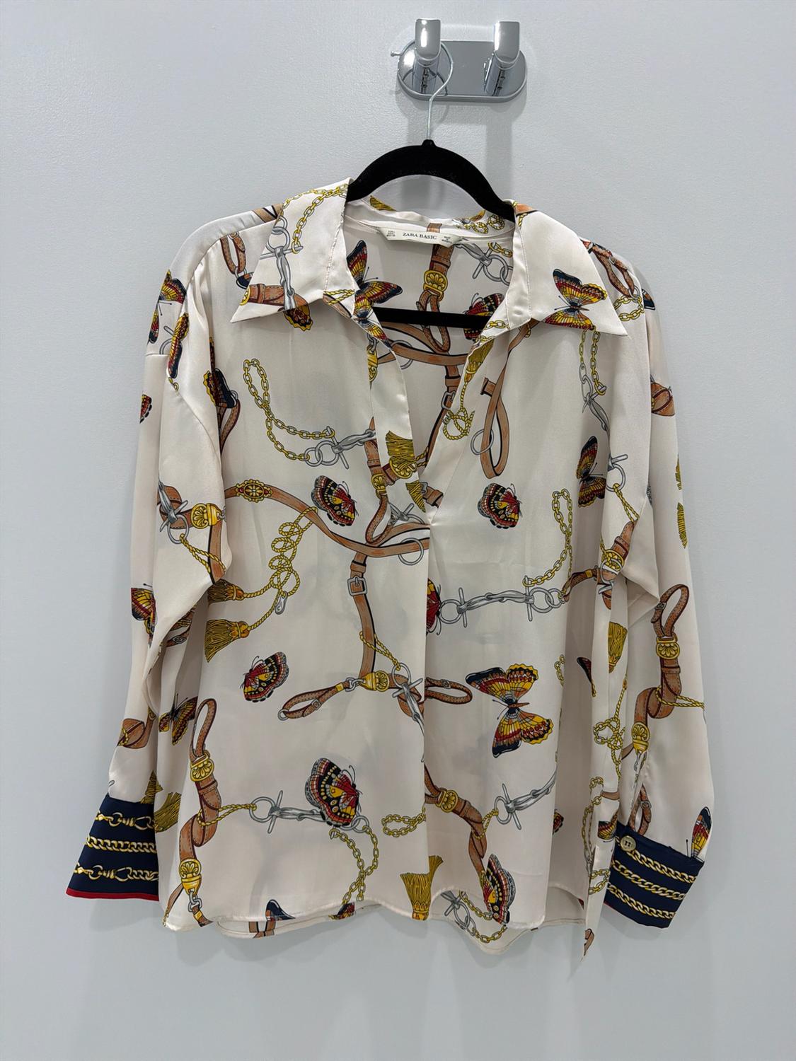 Cream-colored blouse with a print of chains, belts, and butterflies. Dark cuffs. ZARA BASIC label. Hanging on a black hanger against a light background.