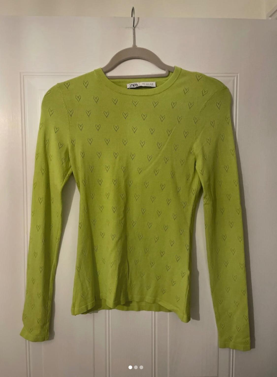 Lime green long-sleeved top with cut-out hearts, hanging on a hanger on a white door. Label with the text ZARA.