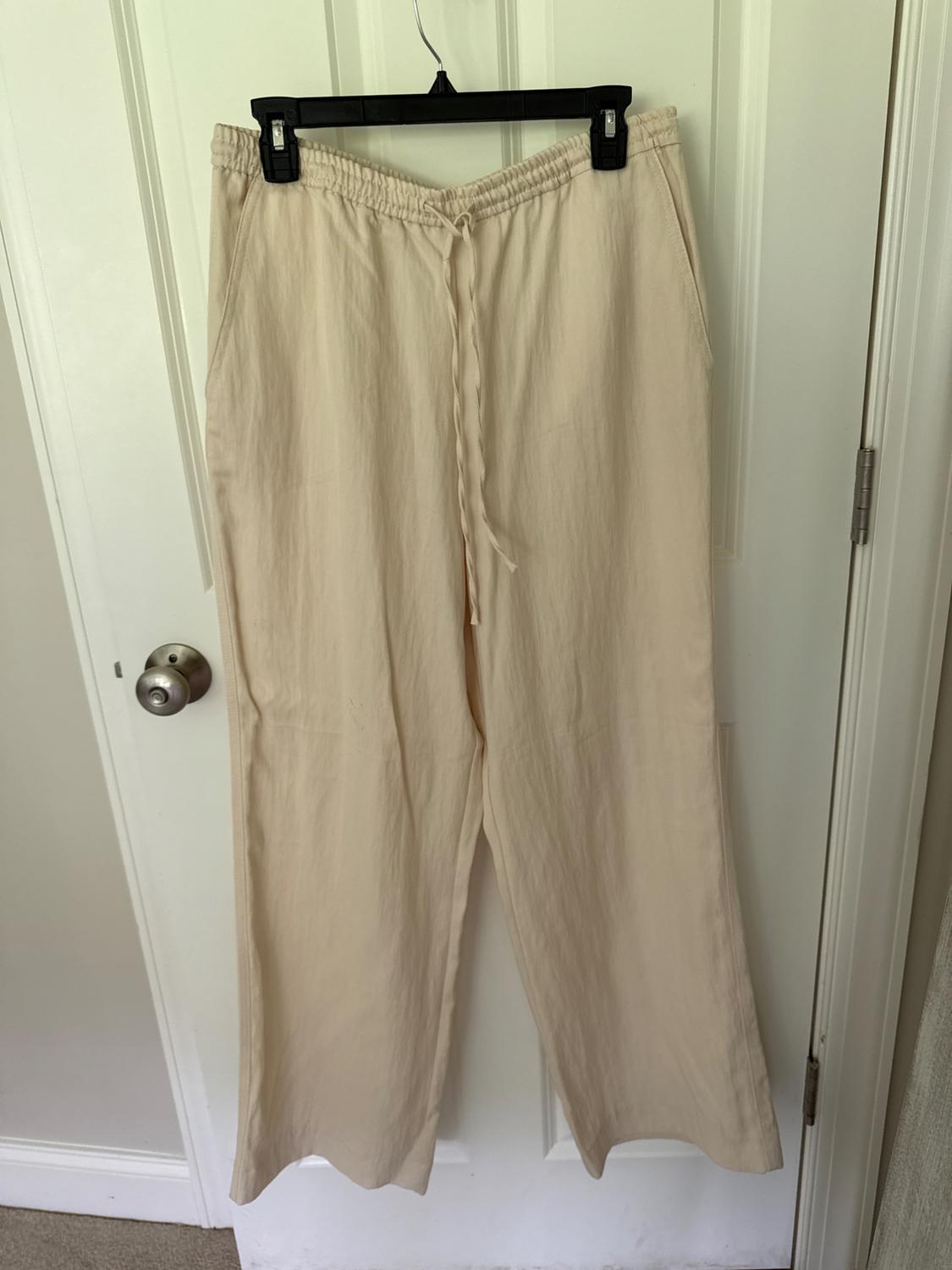 Beige wide-leg pants with an elastic waistband and drawstring, hanging on a black hanger over a white door.