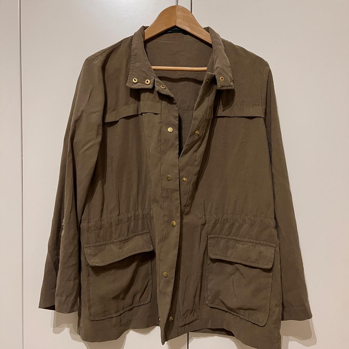 Olive green casual jacket with pockets and gold buttons, hanging on a wooden hanger in front of a white wardrobe. Relaxed style.