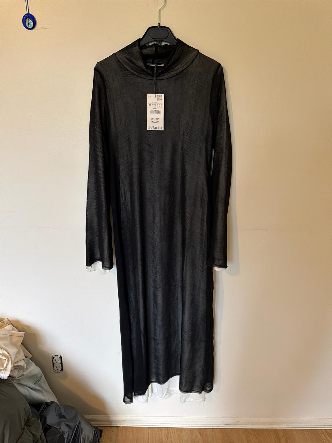 Long-sleeved semi-transparent black maxi dress with tags, hanging on a light-coloured wall. A white garment can be seen underneath.