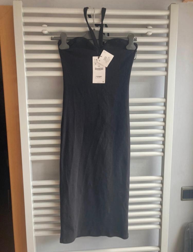 Sleeveless black dress hanging on a hanger over a white radiator. Visible label with product details.