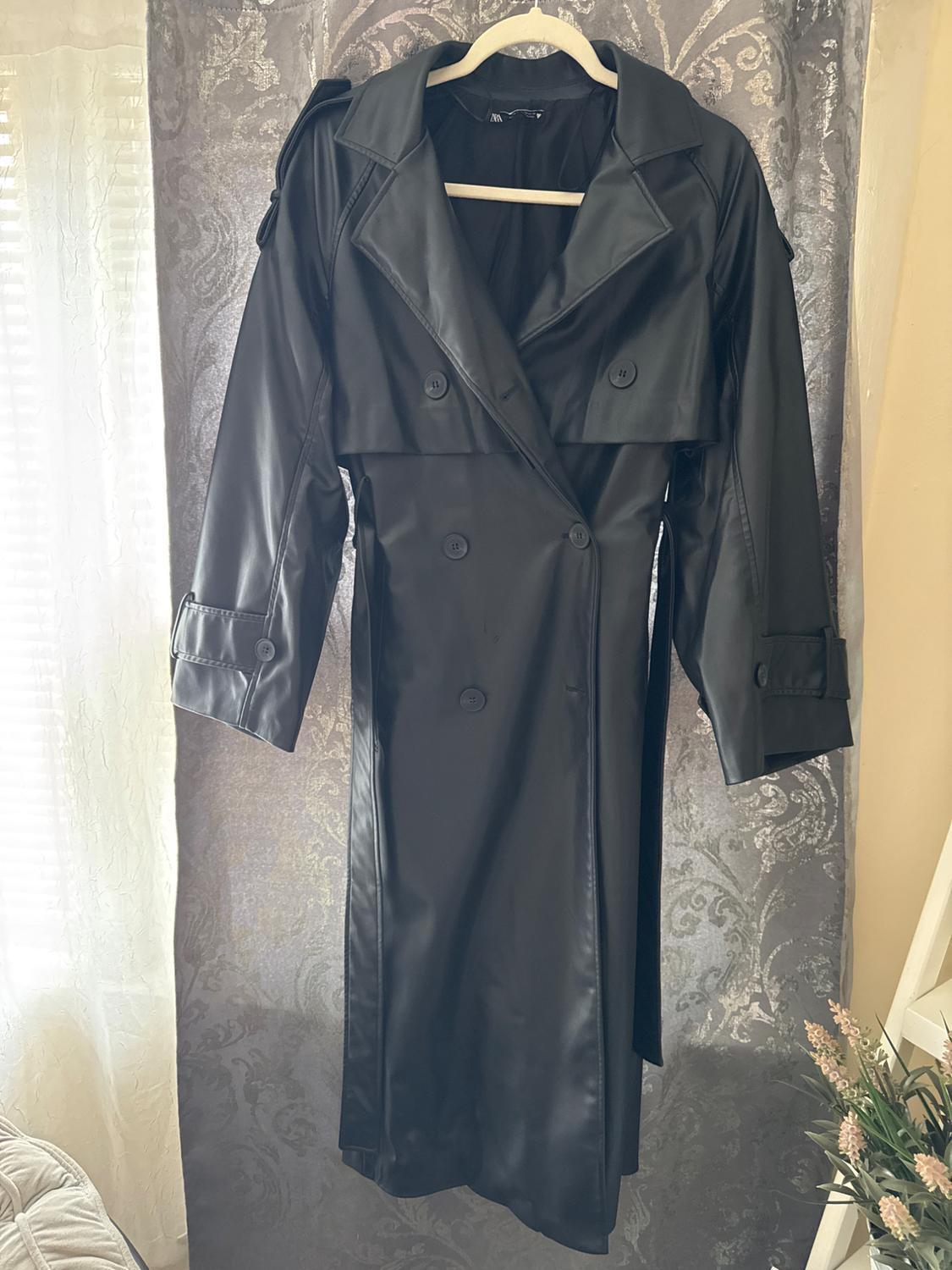 Black trench coat hanging on a hanger against a decorative gray background. Natural light enters through a window on the left.