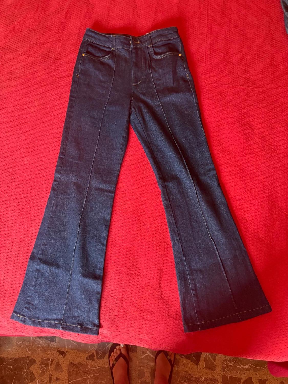Dark blue flared jeans with front seams, laid out on a textured red fabric. Feet are peeking out from the edge.