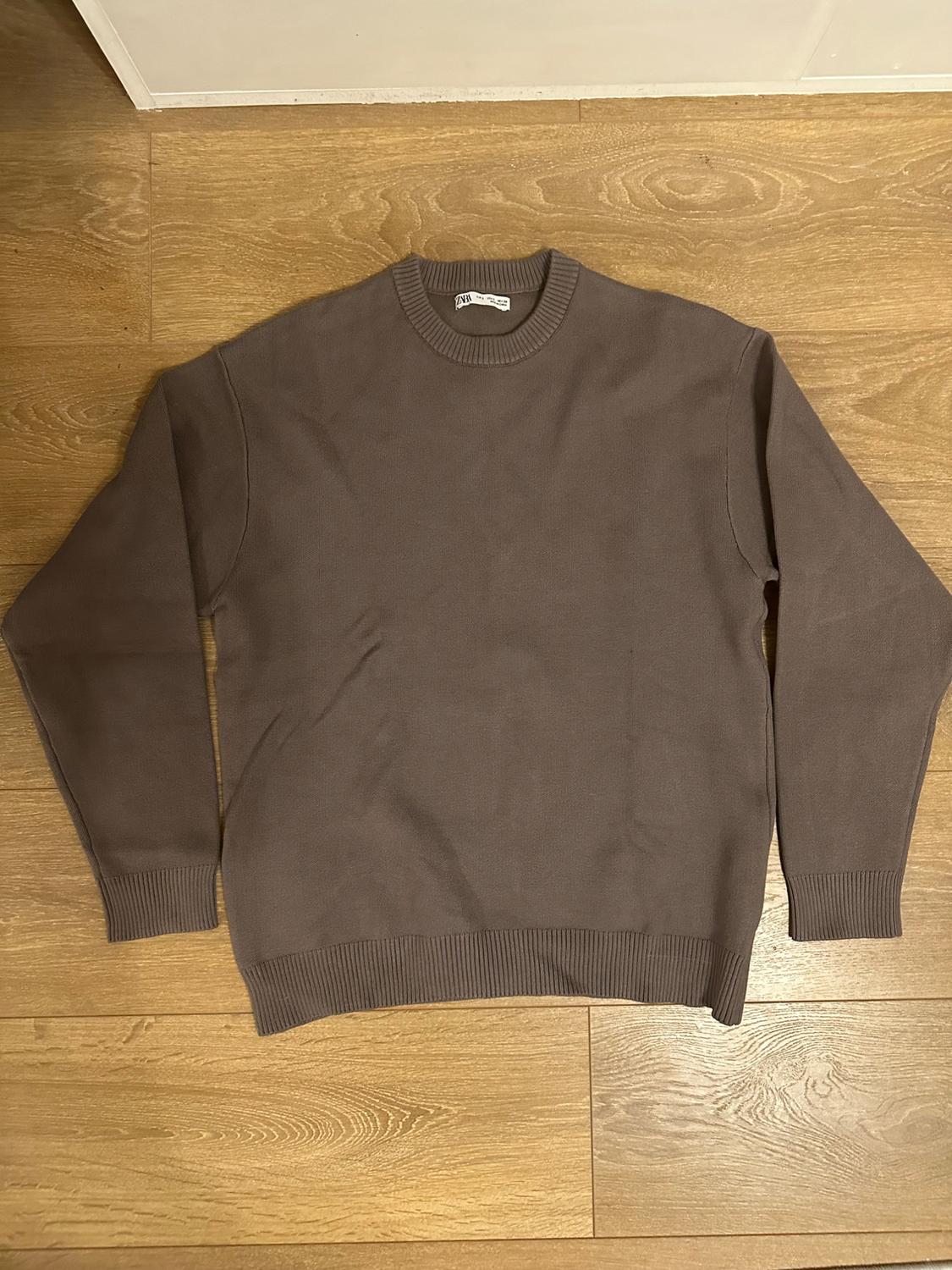 Brown long-sleeved crew-neck jumper, fine knit, on light wooden flooring. Visible label at the neck.