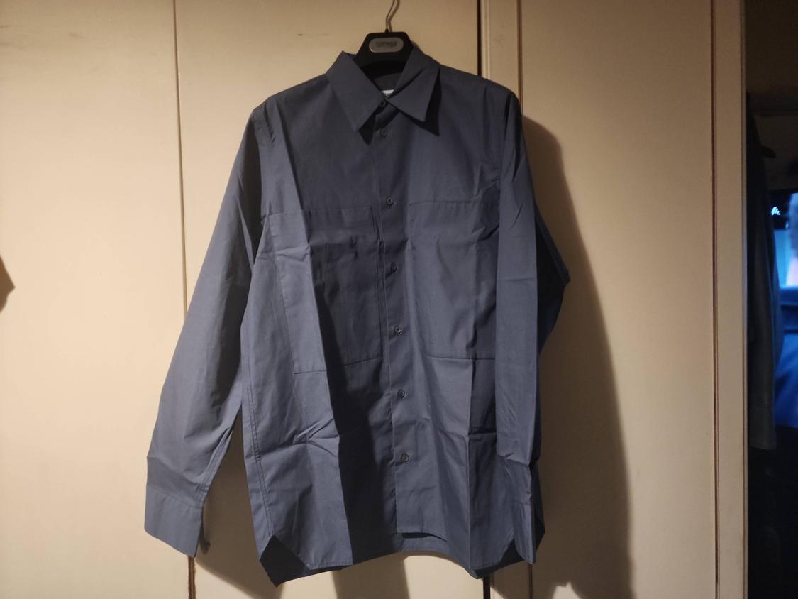 Teal long-sleeved shirt with pleats, hanging on a hanger in front of a light-coloured wardrobe. Casual setting with soft lighting.