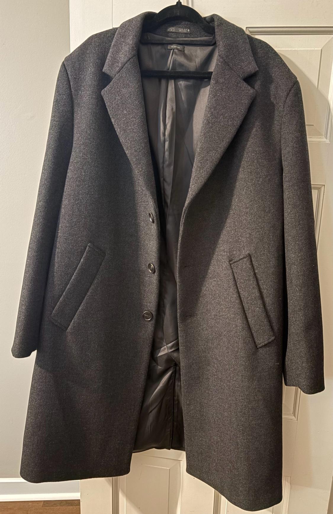 Dark gray long coat with lapels, front buttons, and pockets, hanging on a black hanger on a white door.