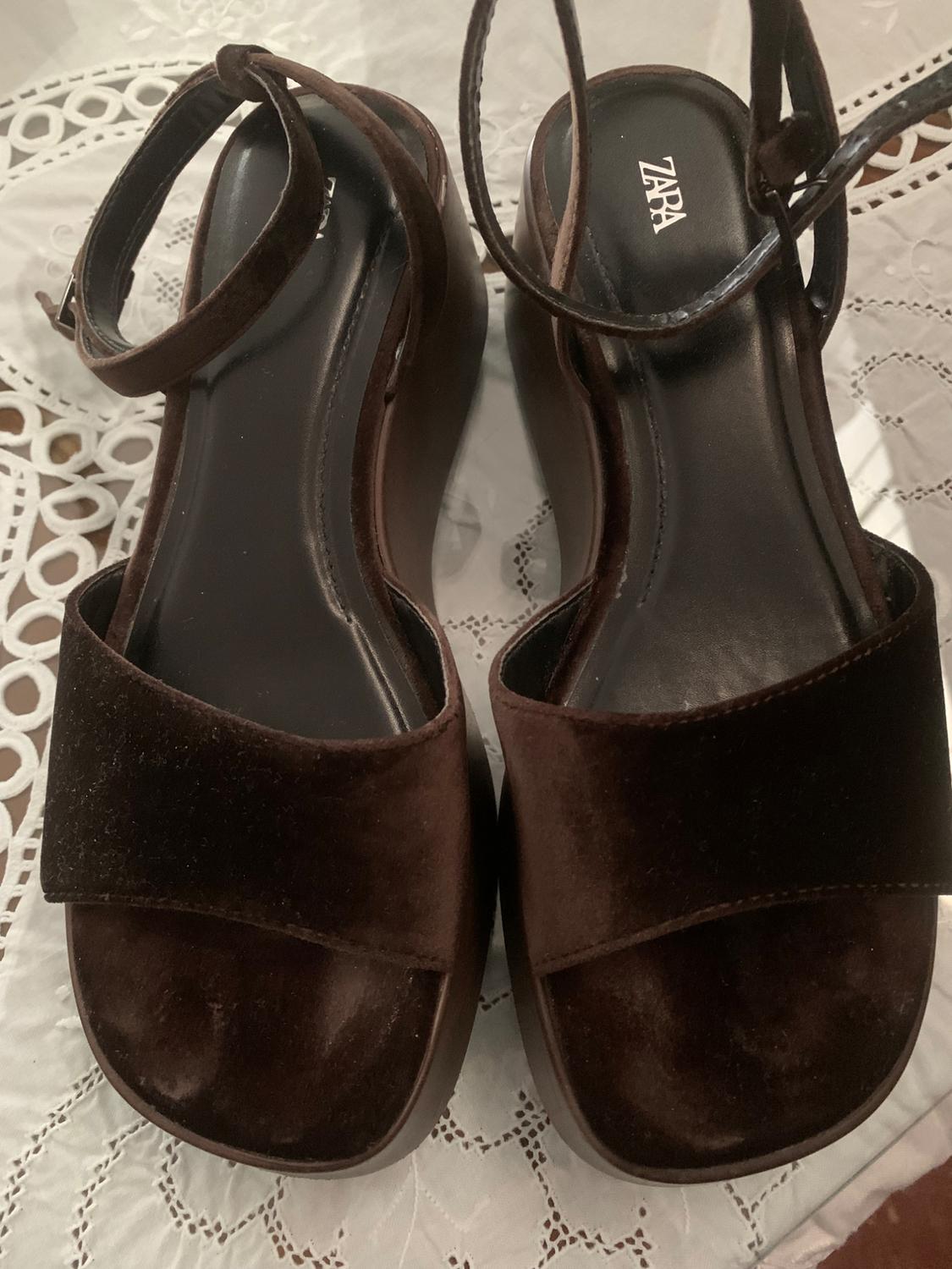 Dark brown platform sandals with adjustable straps, on a white tablecloth with a lace design. Visible brand: Zara.