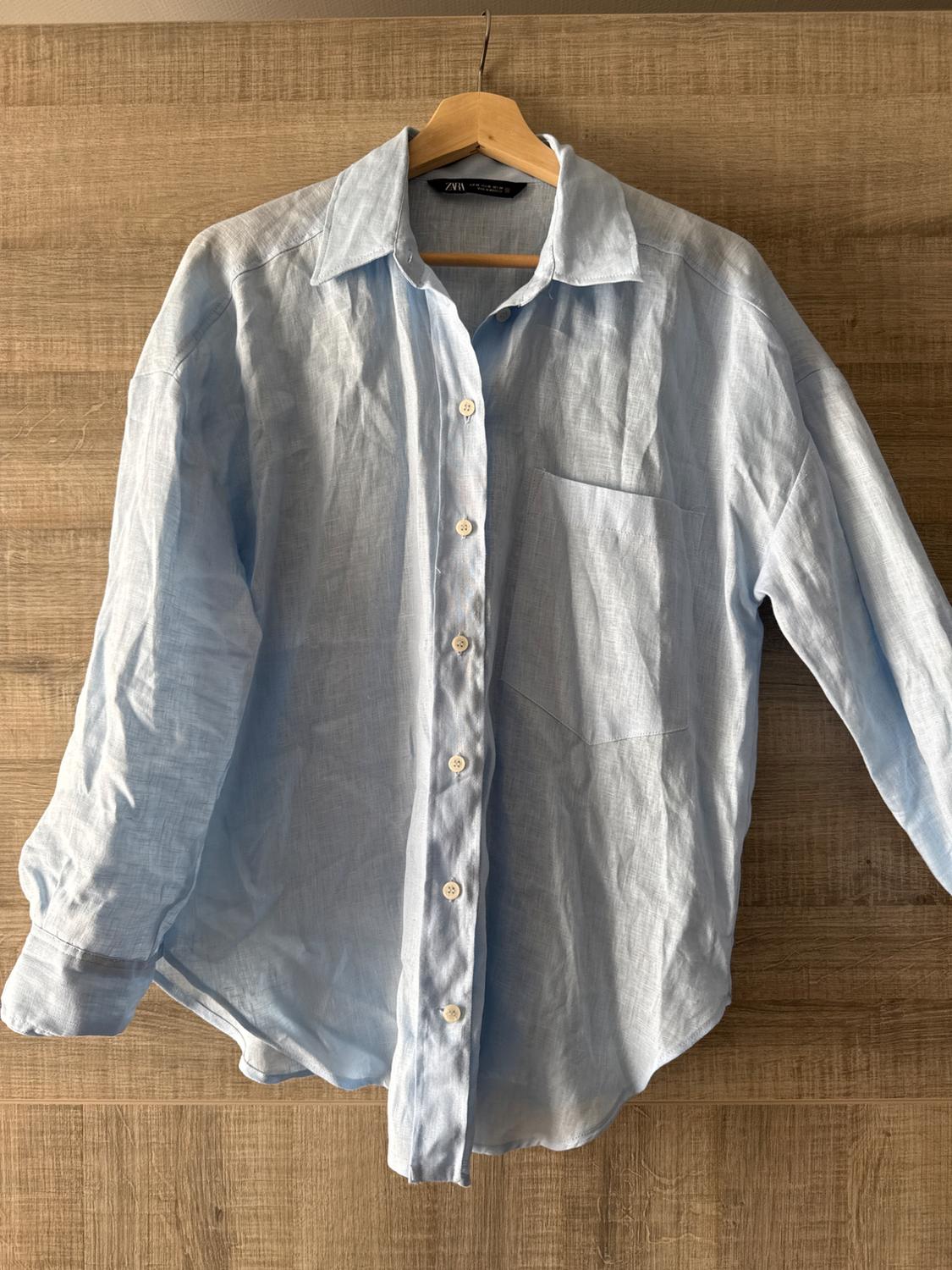 Light blue linen shirt hanging on a wooden hanger against a wooden background. It has white buttons and a chest pocket.