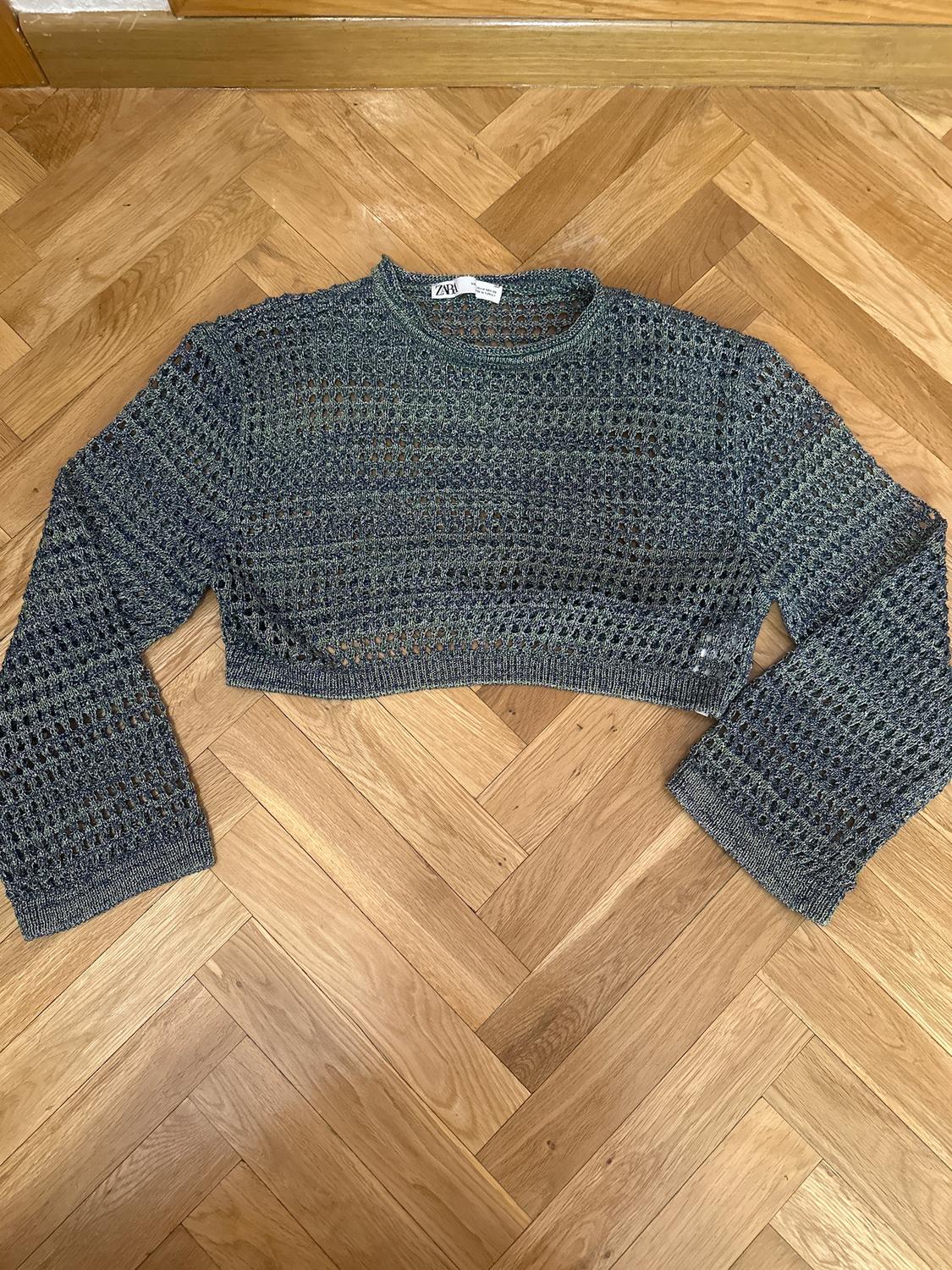 Grey open-knit cropped jumper on a wooden floor. Visible "ZARA" label. Long sleeves and modern design.