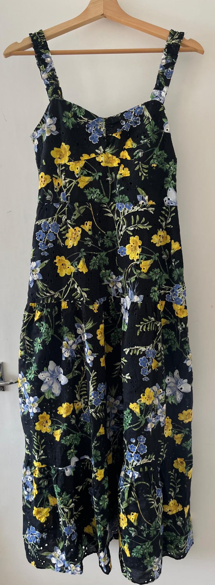Black strappy dress with a yellow and blue floral print and eyelet fabric, hanging on a wooden hanger on a white door.