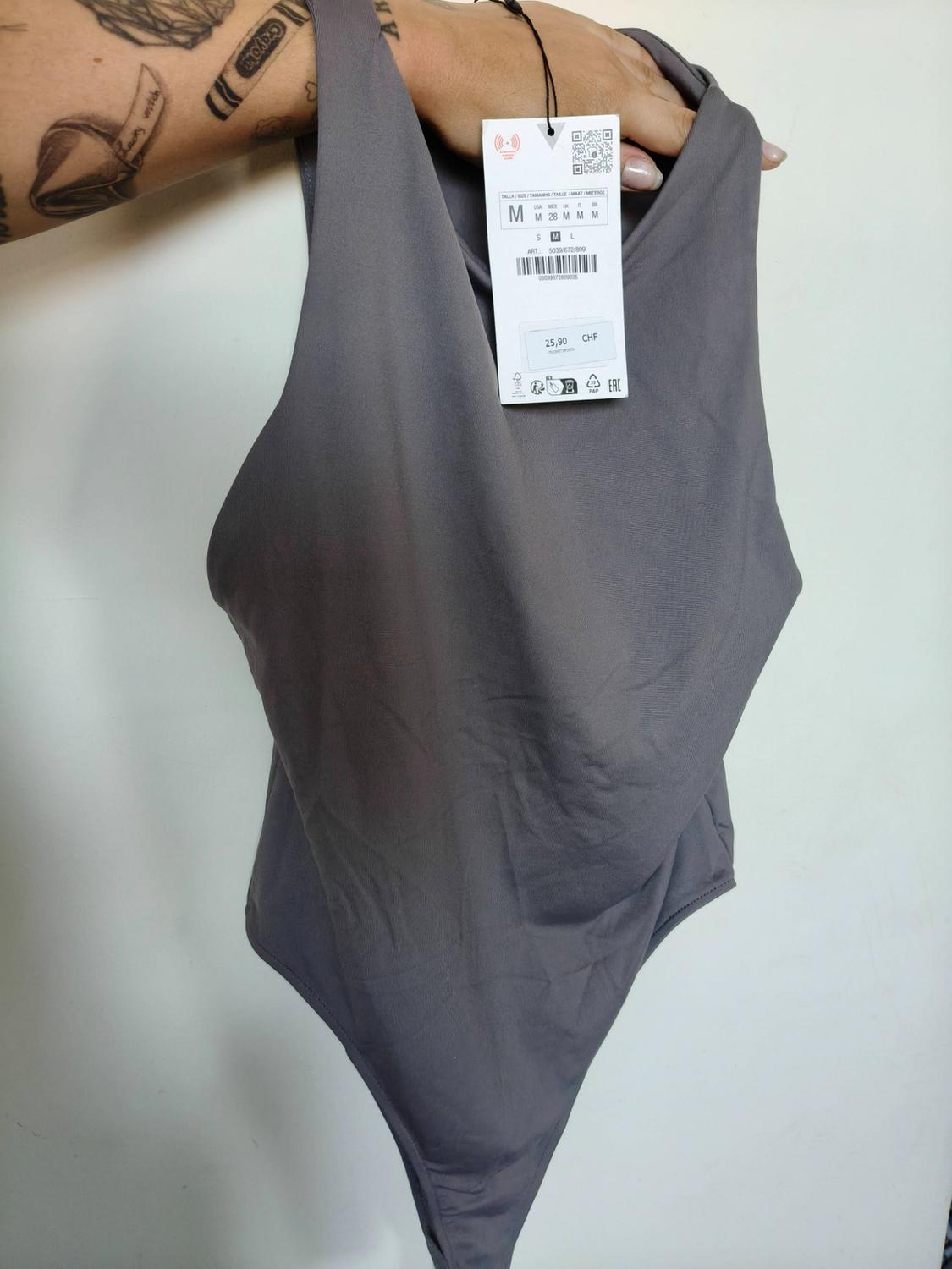 Arm with a Crayola tattoo holding a grey bodysuit against a white background. Label with text: M 28 M M M S M L 25.90 CHF.