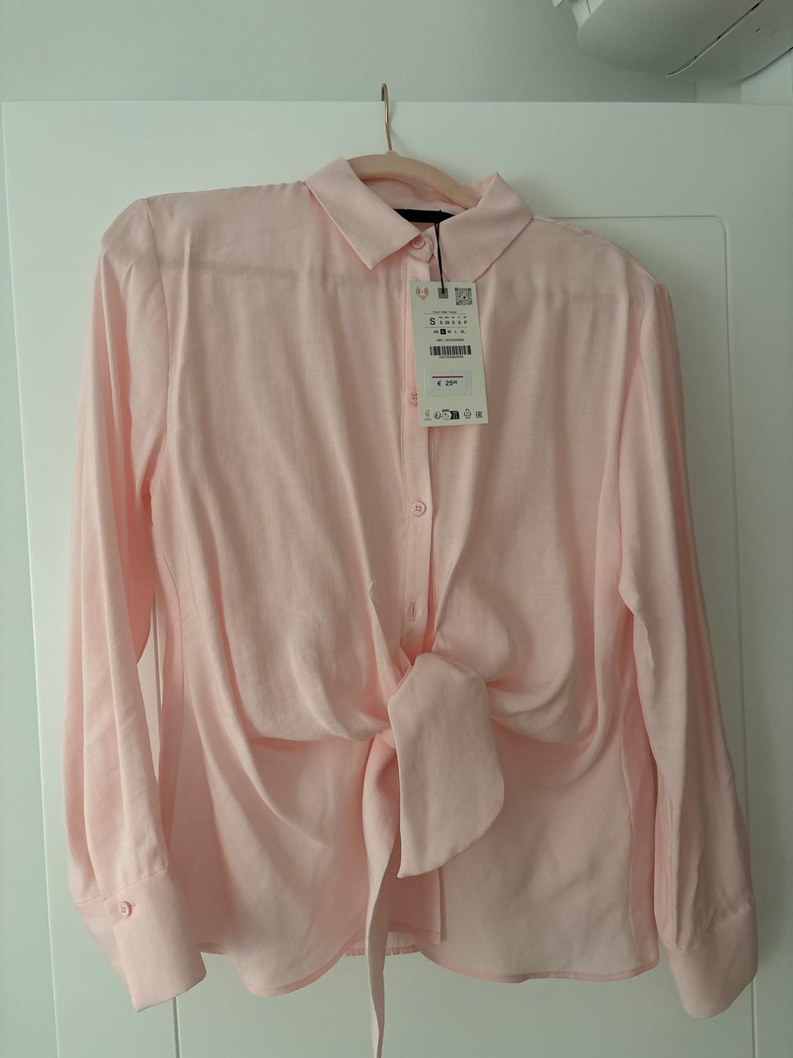 Light pink, long-sleeved blouse with a front knot, hanging on a wooden hanger. Visible tag with price of €29.95.