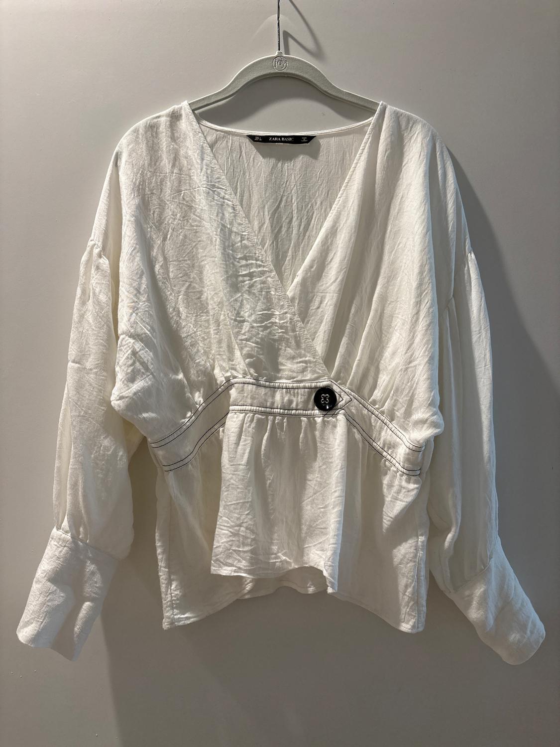 White long-sleeved blouse with a V-neck and a black button at the waist, hanging on a gray hanger against a neutral background.