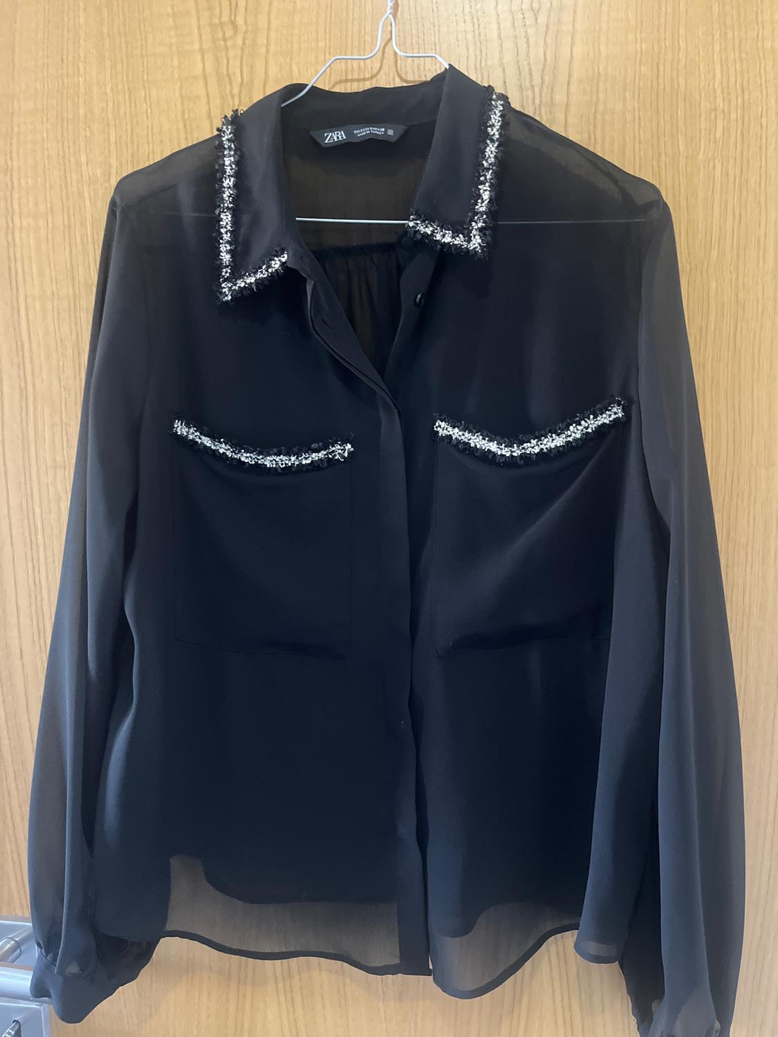 Semi-sheer black blouse with white details on the collar and pockets, hanging on a hanger over a wooden door.