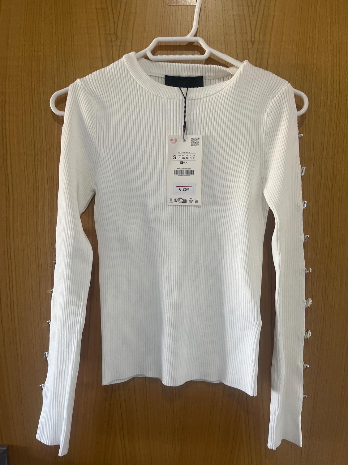 White, long-sleeved jumper with buttons on the sleeves, hanging on a white hanger. Visible tag with price of €25.95.
