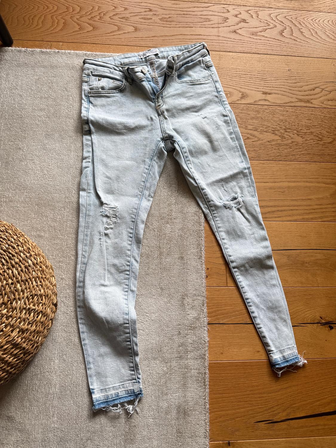 Light-wash skinny jeans with distressed details and a frayed hem, on a beige rug and wooden floor.