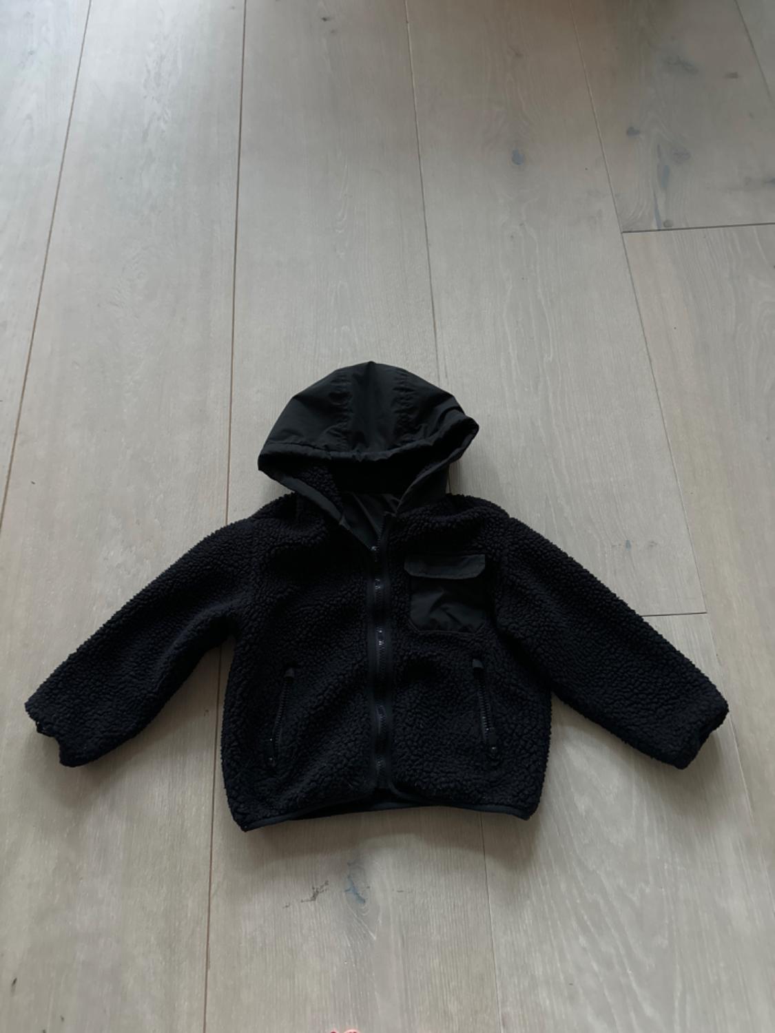 Black wool hooded jacket with zip, on light wooden flooring. Chest pocket and two side pockets.