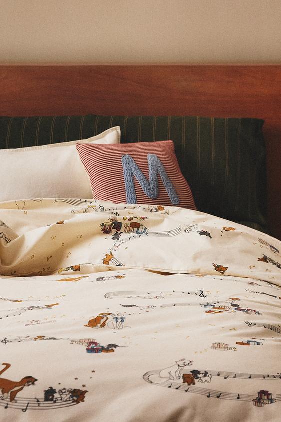 Slatka blagdanska čarolija, kolekcija Zara Home Kids za Božić 6 Posteljina s uzorkom mačaka i notama na krem pozadini, crveni jastuk s plavim slovom "M".