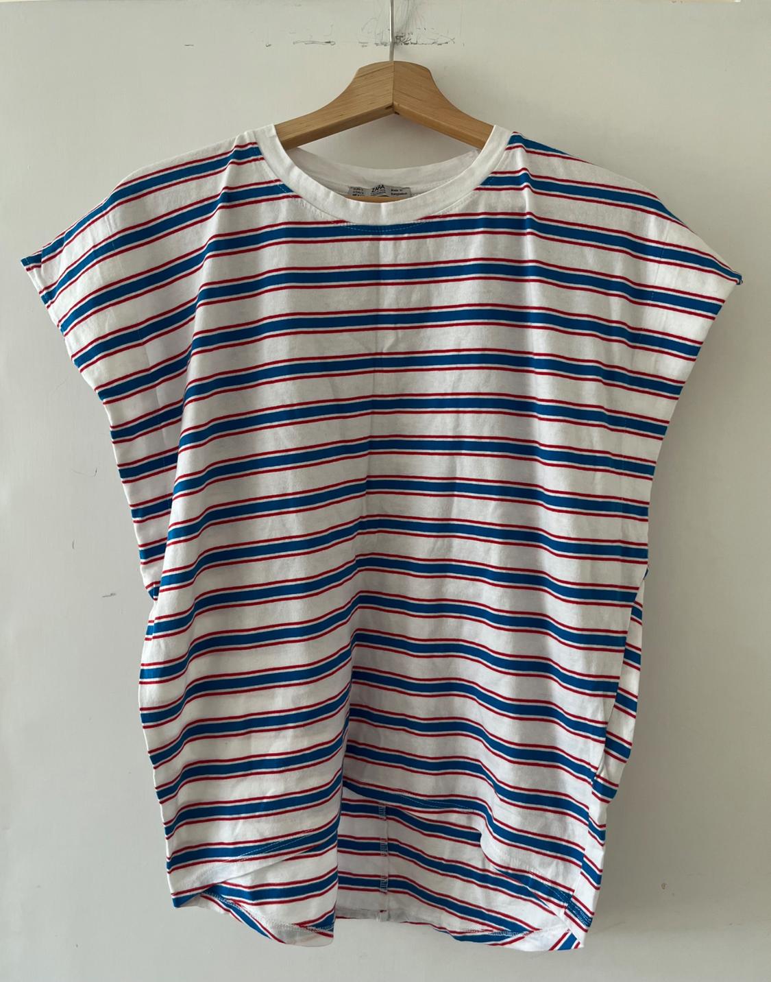 Loose-fitting white short-sleeved T-shirt with blue and red horizontal stripes, hanging on a wooden hanger against a white wall.