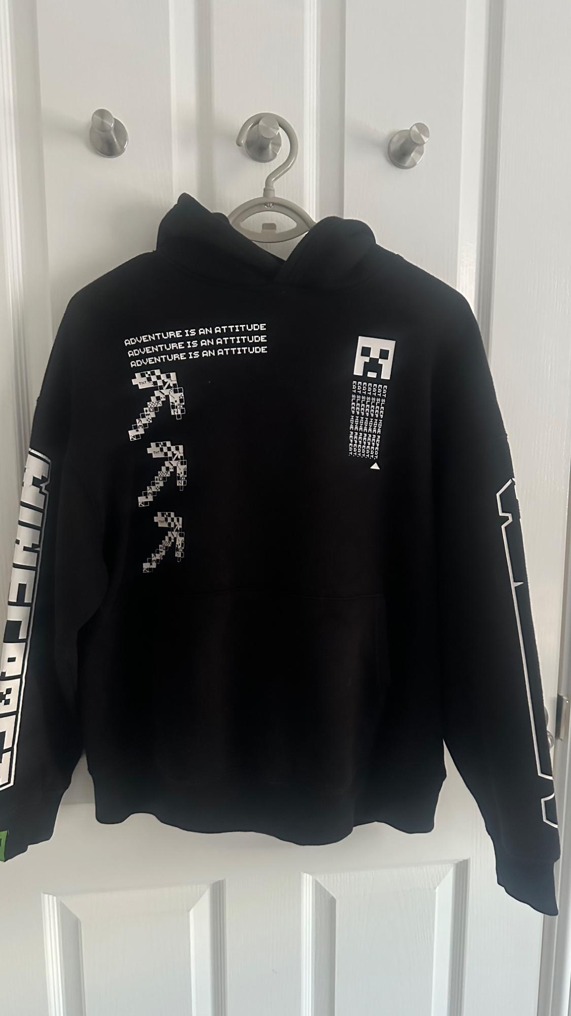 Black hoodie with a hood on a white door. Texts: ADVENTURE IS AN ATTITUDE, EAT SLEEP MINE REPEAT, MINECRAFT and pixelated graphics.