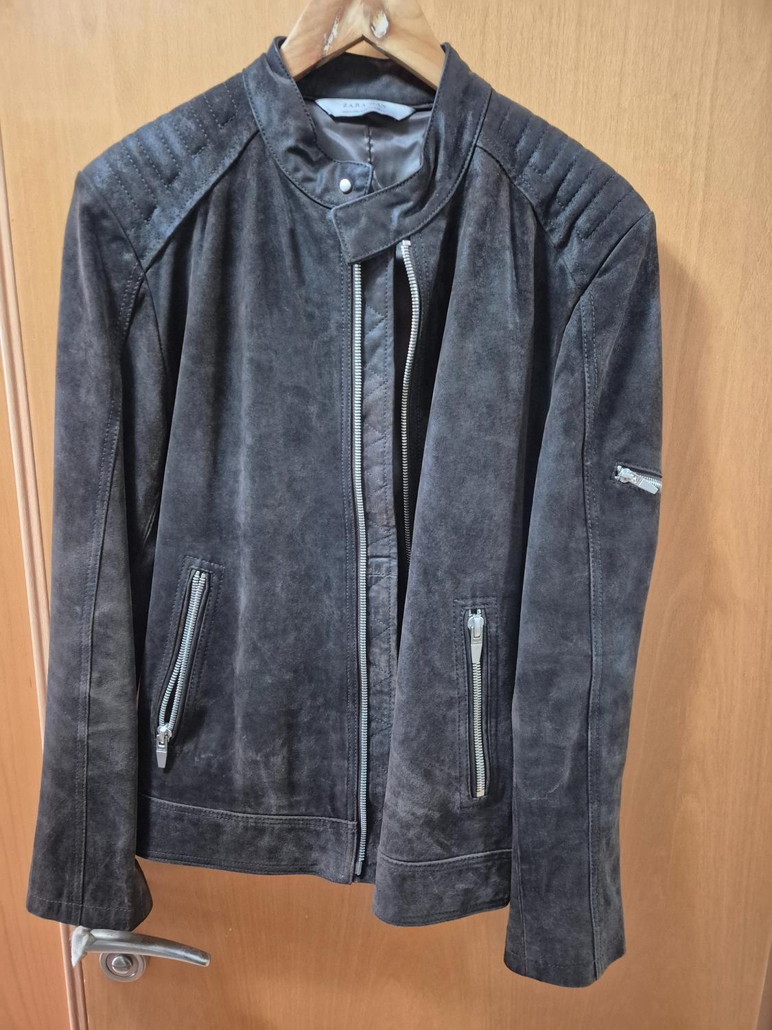 Dark grey suede jacket hanging on a wooden hanger, with silver zips on the pockets and sleeve, over a light wooden door.