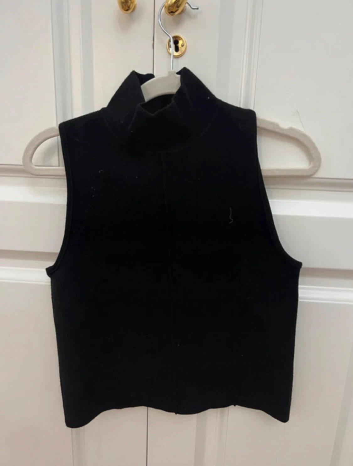 Black sleeveless top, high neck and centre seam, hanging on a hanger on a white door with gold hardware. Casual style.