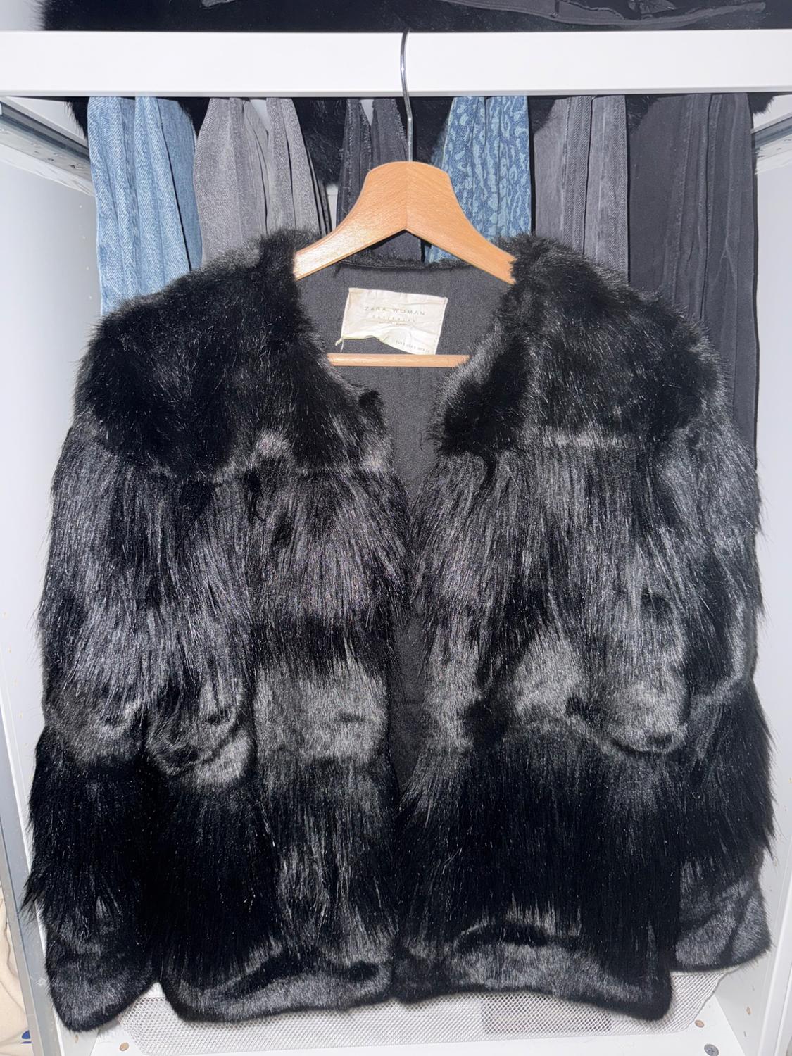 Black faux fur coat hanging on a wooden hanger, with a background of different coloured garments in a wardrobe.
