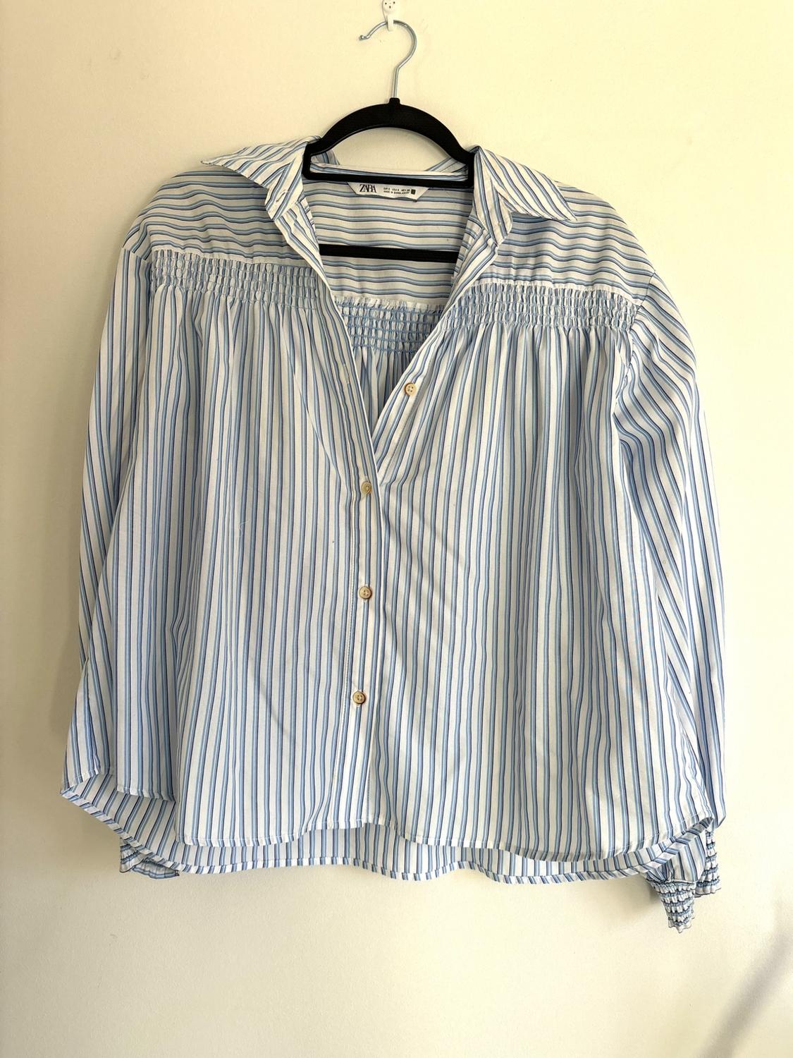 Blue and white striped shirt hanging on a black hanger. It has gold buttons and a ruched design on the chest.