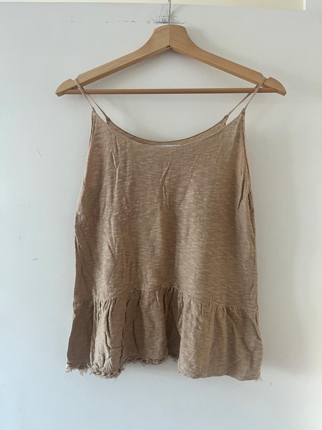 Beige camisole top with spaghetti straps, featuring a frilled hem and frayed edges, hanging on a wooden hanger against a white wall.