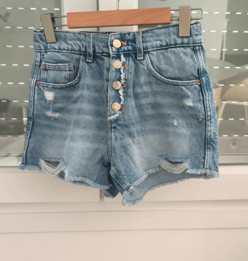 Light-blue denim shorts hanging on a hanger. They feature a high waist, a front button fastening, distressed details and a frayed hem.