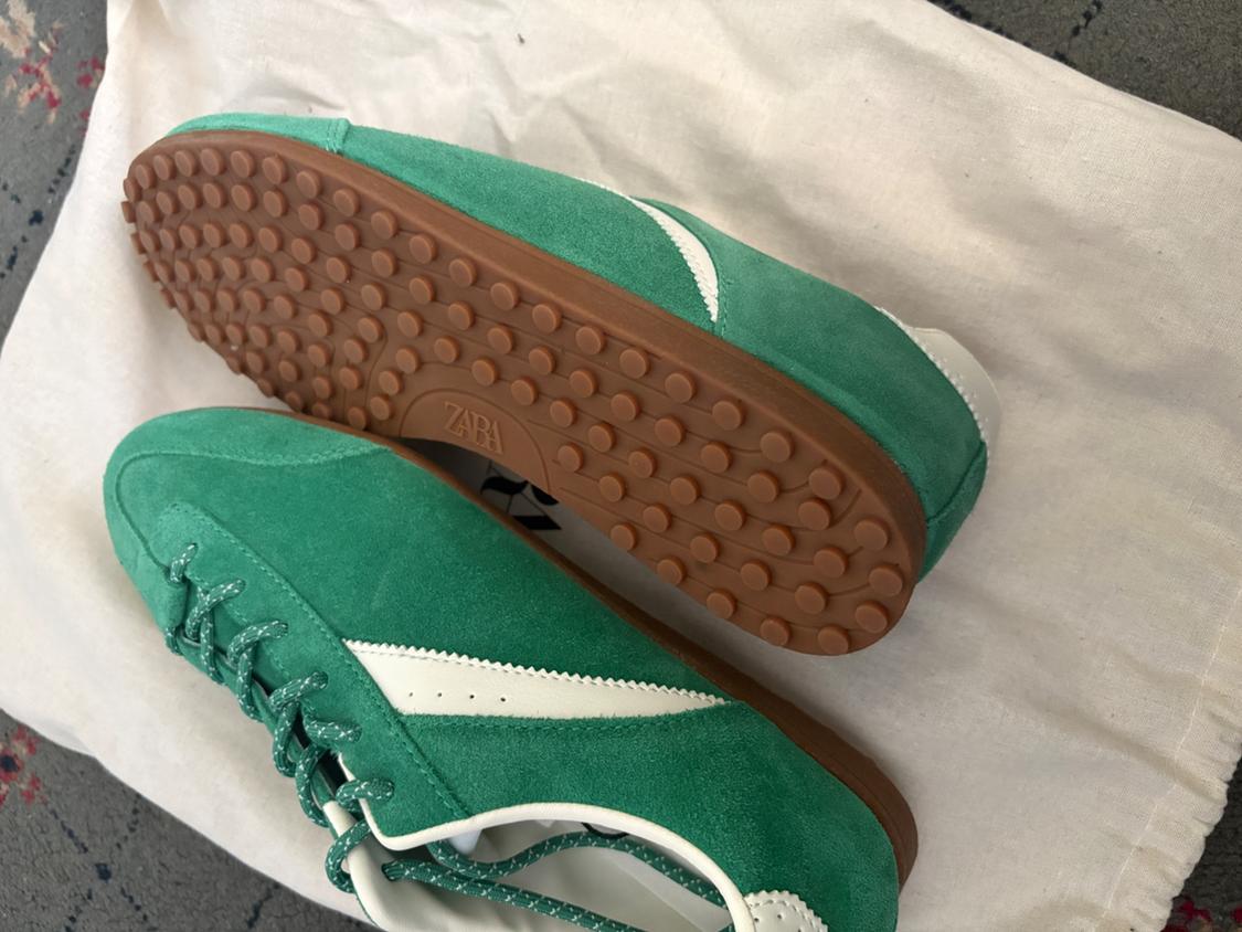 Green suede trainers with white details on a white fabric background. One shows the brown rubber sole with a dotted texture and the word ZARA.