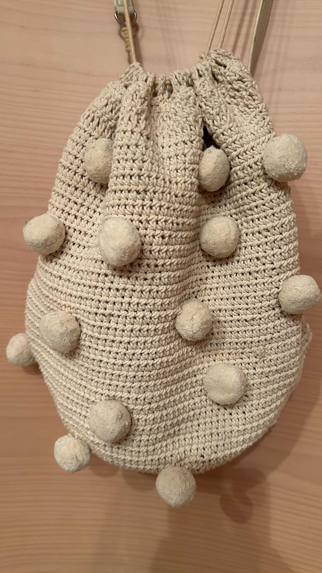 Beige crochet bucket bag with decorative pom-poms, hanging against a light wood background.