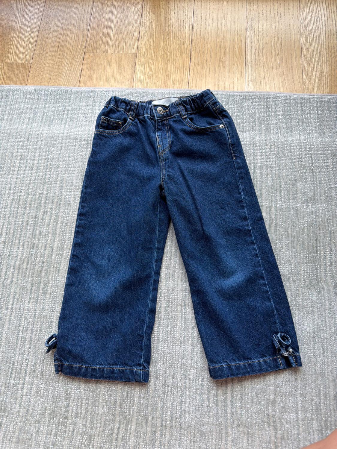 Blue denim jeans for children with an elasticated waist and ties at the cuffs, on a grey rug and wooden floor.